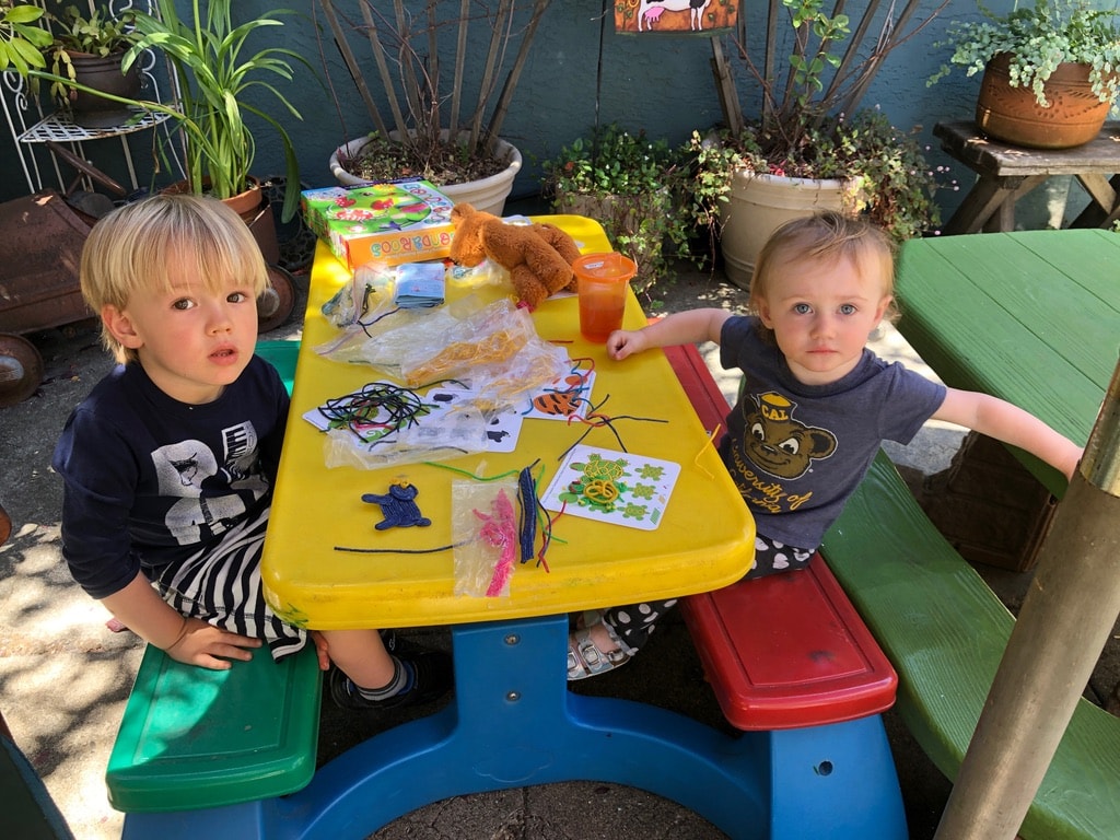 Two toddlers crafting together at a yellow table outdoors